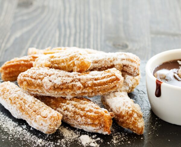 Crispy churros heavily dusted with sugar, piled on a slate surface with a side of melted chocolate, perfect for dipping, on a rustic wooden background.
