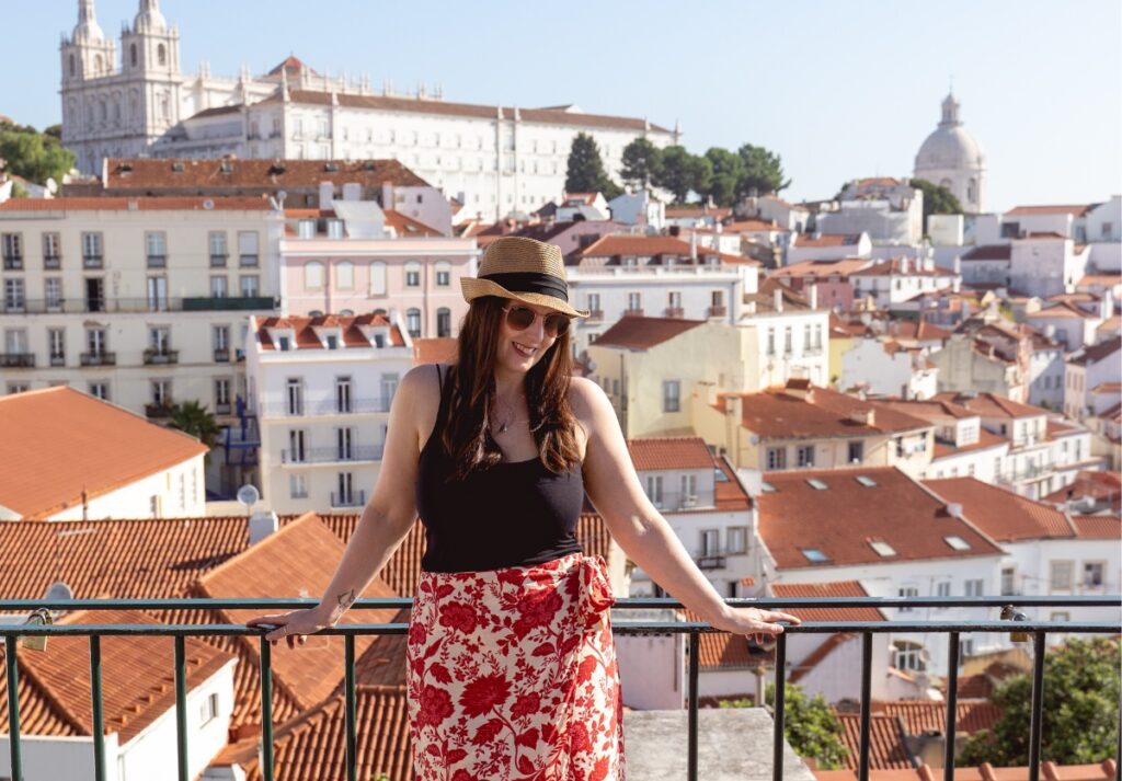 A smiling woman leans on a balcony railing with a panoramic view of Lisbon, Portugal, in the background. She wears a summer outfit with a black top and a red floral skirt, complemented by a stylish straw hat and sunglasses. Behind her, the terracotta rooftops of Lisbon's buildings spread out towards the historic São Vicente de Fora monastery and the National Pantheon, visible under a clear blue sky.