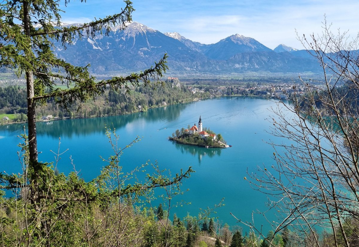 A breathtaking aerial view of Lake Bled in Slovenia, with its iconic church on an island in the center of the emerald-green lake. Snow-capped mountains loom in the background, framing the tranquil water and lush greenery surrounding the lake's perimeter.