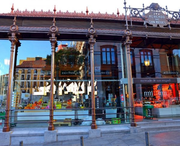 The elegant facade of Mercado San Miguel in Madrid, featuring ornate ironwork and glass panels, with the vibrant display of market goods visible through the windows, set against the clear blue sky of a sunny day.