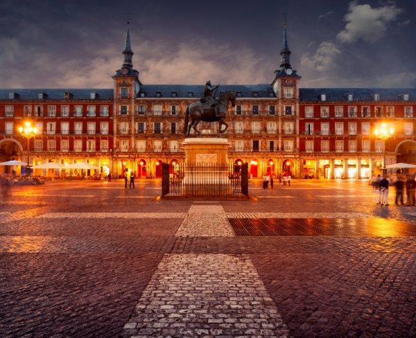 The historic Plaza Mayor of Madrid at dusk, illuminated by warm lights, featuring the iconic equestrian statue of King Philip III and the plaza's traditional Spanish architecture, under a dramatic sky.