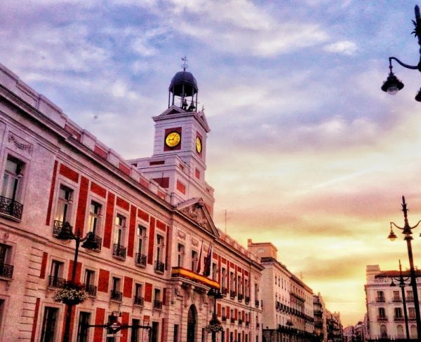 The Puerta del Sol in Madrid, featuring the iconic red and white façade of the Casa de Correos with its clock tower, bathed in the rosy hues of sunset, and the street lights beginning to glow as evening approaches.