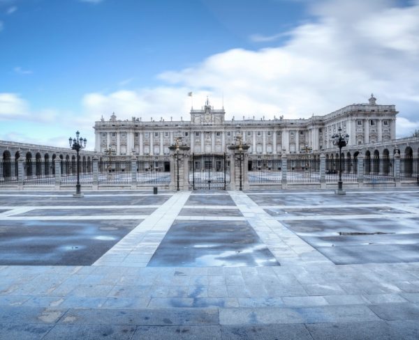 The Royal Palace of Madrid captured under a cloudy sky, showcasing its majestic architecture and grandeur, with the empty foreground of the plaza's patterned pavement reflecting the building's facade.
