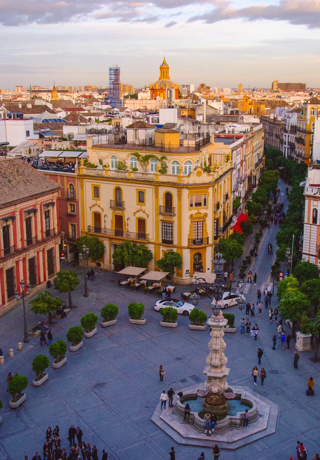 This is a vibrant, high-angle view of a bustling square in Seville, Spain, during the early evening. The square is framed by classic European buildings with pastel facades; one prominently in a sunny shade of yellow with intricate architectural details. People appear as tiny figures, walking and congregating around the central fountain, which is a classic tiered design with water basins. The streets are lined with trees and outdoor café seating, inviting social interaction. Beyond the square, the densely packed houses and buildings stretch into the distance, with the city's iconic rooftops and church spires under a soft, pastel-colored sky.
