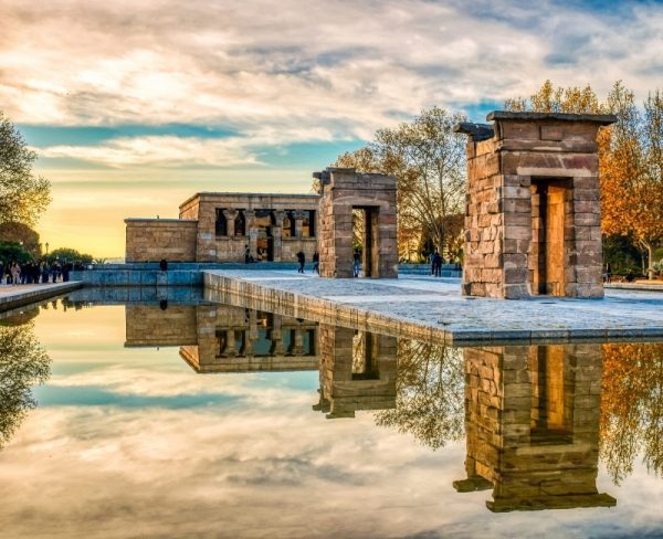 The Temple of Debod in Madrid, an ancient Egyptian temple set against a reflective pool, captured at sunset with golden light accentuating the stone structure and autumnal trees.