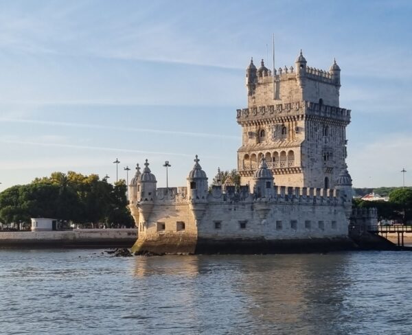 A view of the historic Belém Tower in Lisbon, seen from the water. The tower's ornate, fortress-like architecture stands prominently against the backdrop of a clear sky and nearby greenery. The water in the foreground reflects the tower, enhancing its majestic appearance.