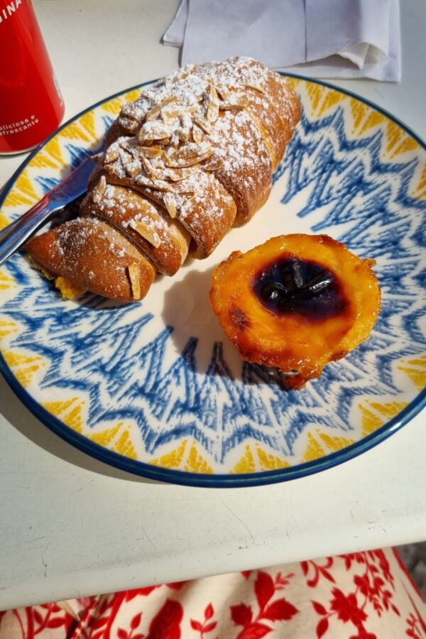 A plate with a gluten-free pastel de nata and an almond croissant, dusted with powdered sugar. The plate features a vibrant blue and yellow pattern, adding a cheerful touch to the presentation. In the background, part of a red and white floral fabric is visible