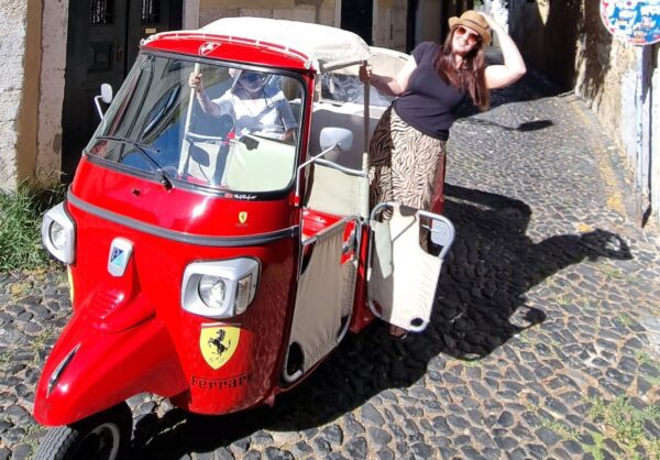 A woman in a straw hat and sunglasses is leaning out of a red tuk-tuk on the cobbled streets of Lisbon. The tuk-tuk, with a Ferrari logo on its side, is driven by a person in the front seat. The scene captures a fun and adventurous moment in the historic streets of the city.