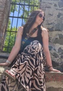 Me wearing sunglasses, a black tank top, and a zebra-print skirt is sitting on a stone structure. The background features a window with metal bars and green foliage visible through it. She is smiling and looking to the side, enjoying a sunny day outdoors.