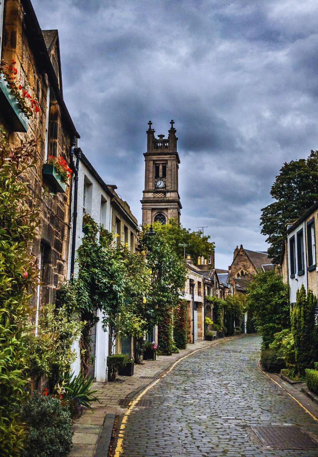 A narrow, cobblestone street in Scotland, lined with charming old stone buildings adorned with greenery and flowers, leads towards a tall, historic clock tower under a moody, cloudy sky.