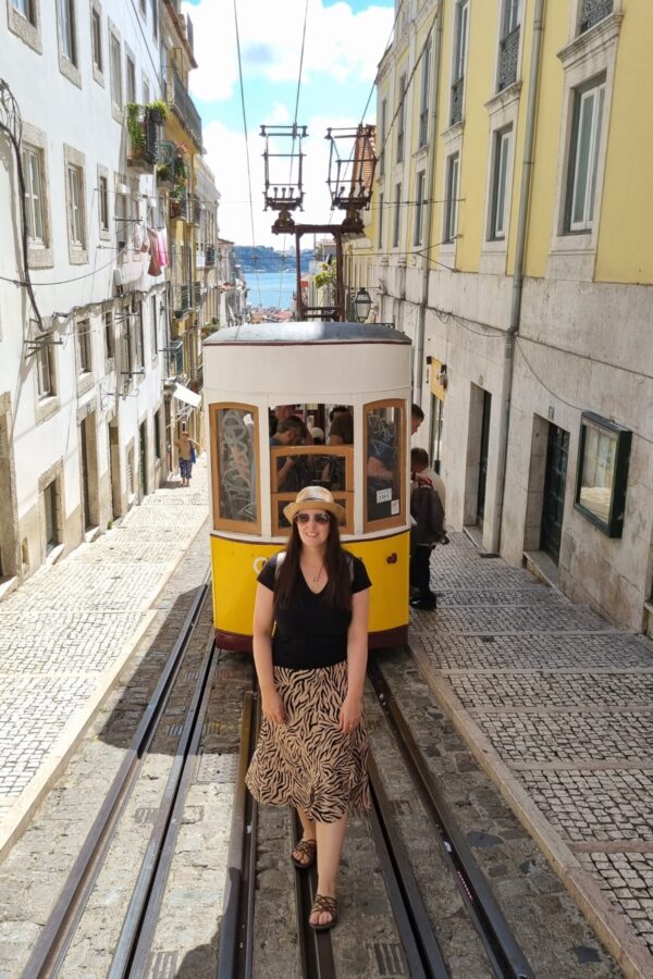 A woman wearing a straw hat and sunglasses stands confidently on the tracks in front of a yellow funicular in Lisbon. The funicular is making its way down a steep, narrow street lined with historic buildings, with the Tagus River visible in the background. The scene captures a moment of exploration and urban charm in the heart of the city.
