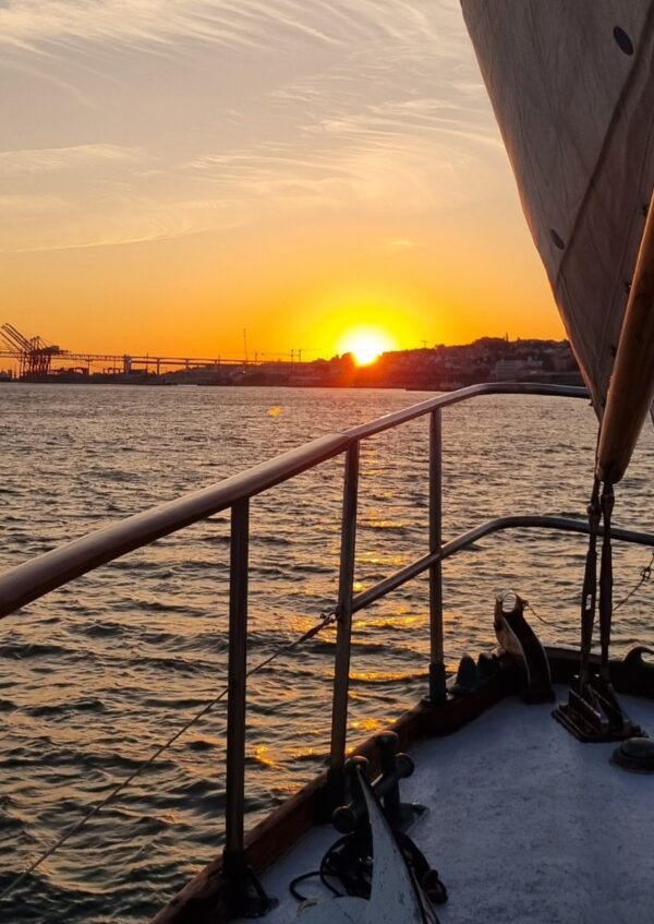 A breathtaking sunset view from a wooden sailboat, with the sun setting over the horizon and casting a warm glow over the water. The boat's railing and sail are visible, with distant silhouettes of cranes and buildings adding to the serene, picturesque scene.