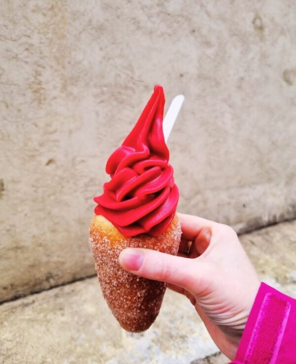 The image captures a hand holding a chimney cake filled with vibrant red soft-serve ice cream against a plain background. The chimney cake is sugar-coated, and the ice cream forms a tall, swirled peak with a small spoon inserted.