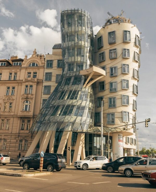 The image features the Dancing House in Prague, an iconic modern architectural structure designed by Frank Gehry and Vlado Milunić. The building has a dynamic, twisted design with one tower resembling a glass dress, giving the appearance of two dancers. It is set against a backdrop of traditional architecture and a partly cloudy sky, with cars passing by in the foreground.