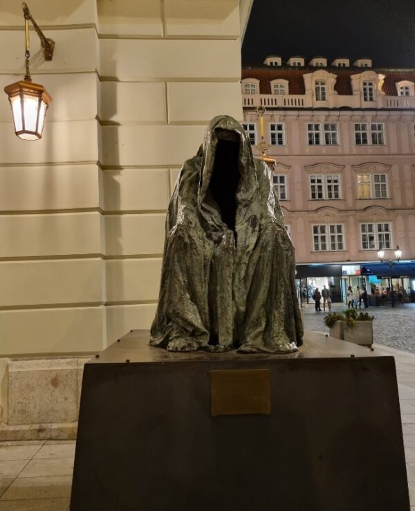 This image shows a cloaked, hooded statue known as "Il Commendatore" by Anna Chromy, positioned against a building with warm lighting. The statue has an eerie, empty hood and draped robes, creating a mysterious and ghostly appearance.