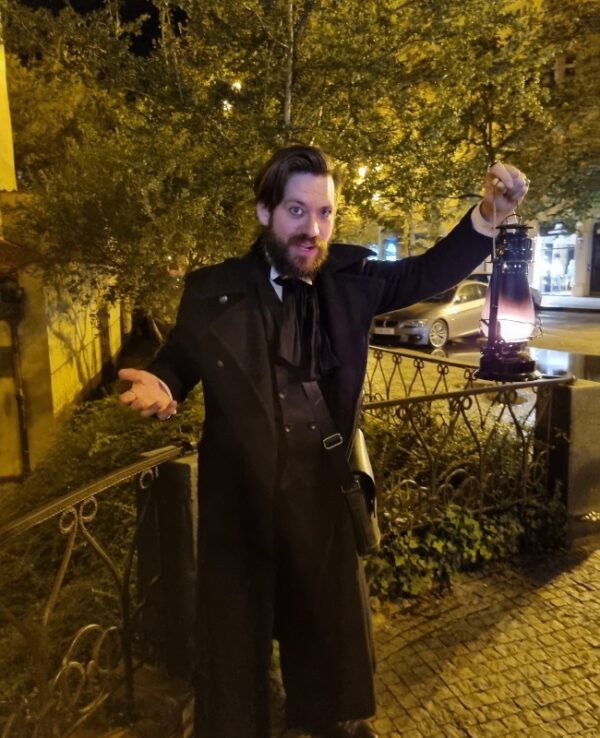 This image features a man dressed in a historical costume, holding a lantern during a ghost tour in Prague. He has a dramatic expression and gestures, set against a nighttime backdrop with warm streetlights and surrounding foliage.