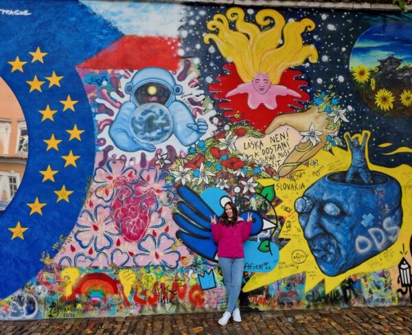 The image shows a person standing in front of the colorful John Lennon Wall in Prague. The wall is covered in vibrant graffiti and murals, including a depiction of an astronaut holding the Earth, a golden-haired figure, and various symbols and messages. The person is wearing a bright pink jacket, blending into the vivid background.