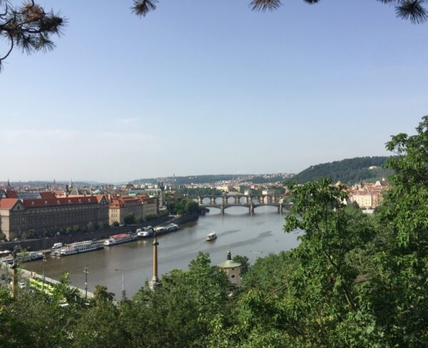 The image offers a panoramic view of Prague from Letná Park, overlooking the Vltava River and multiple historic bridges. The red rooftops of buildings and lush greenery frame the scene, with boats visible on the river under a clear, sunny sky.