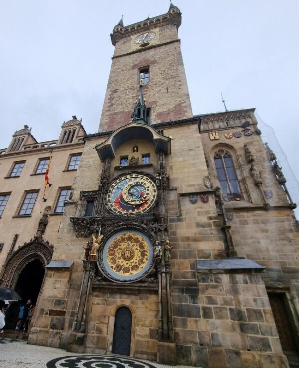 The image depicts the Prague Astronomical Clock, also known as the Orloj, mounted on the Old Town Hall in Prague. The clock features intricate details, including a large dial with astrological symbols and a calendar dial below it, set against a historic stone building.