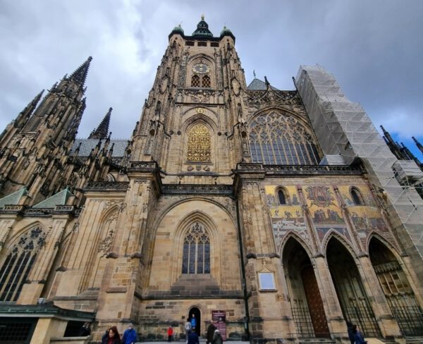 The image captures the grand facade of St. Vitus Cathedral within Prague Castle. The gothic architecture features intricate details, large stained glass windows, and ornate gold accents. Part of the structure is covered in scaffolding, indicating ongoing restoration work, with people walking near the entrance.