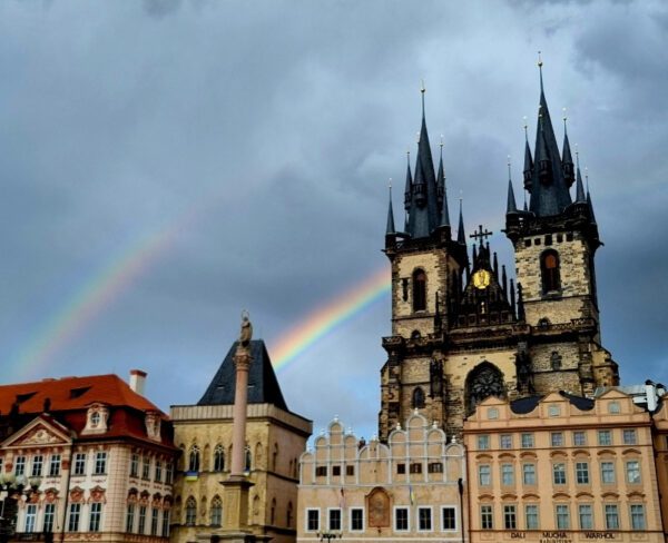 The image shows the Church of Our Lady before Týn in Prague’s Old Town Square, characterized by its twin gothic spires. A vibrant double rainbow arches over the historic buildings, contrasting with the dramatic, cloudy sky.