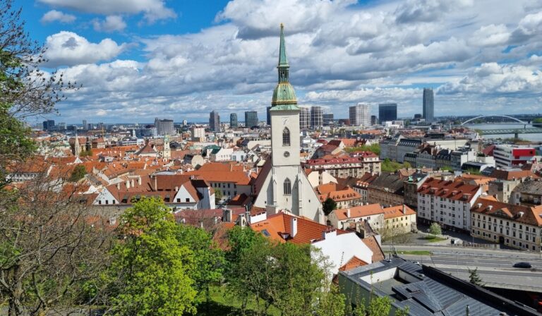 Panoramic view of Bratislava, Slovakia, featuring the prominent St. Martin’s Cathedral with its green spire rising above red-roofed buildings. The scene blends the historic Old Town in the foreground with modern high-rises and the UFO Bridge visible in the distance under a partly cloudy sky.