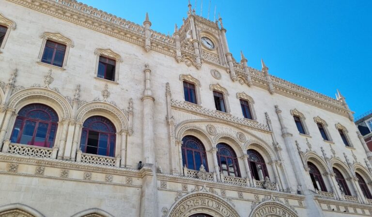 This image shows the ornate façade of the Rossio Railway Station in Lisbon, Portugal. The building features neo-Manueline architecture with intricate stonework, arched windows framed by detailed carvings, and a central clock above the main entrance. The bright blue sky contrasts beautifully with the light-colored stone, highlighting the structure’s historic and decorative charm.