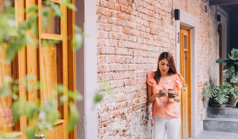 A young woman leans against a rustic brick wall outside a building with wooden doors, holding a coffee cup in one hand and looking at her phone in the other. She’s wearing a peach blouse with ruffled sleeves and white pants, standing in a quiet, plant-lined walkway that gives off a cozy, urban vibe.