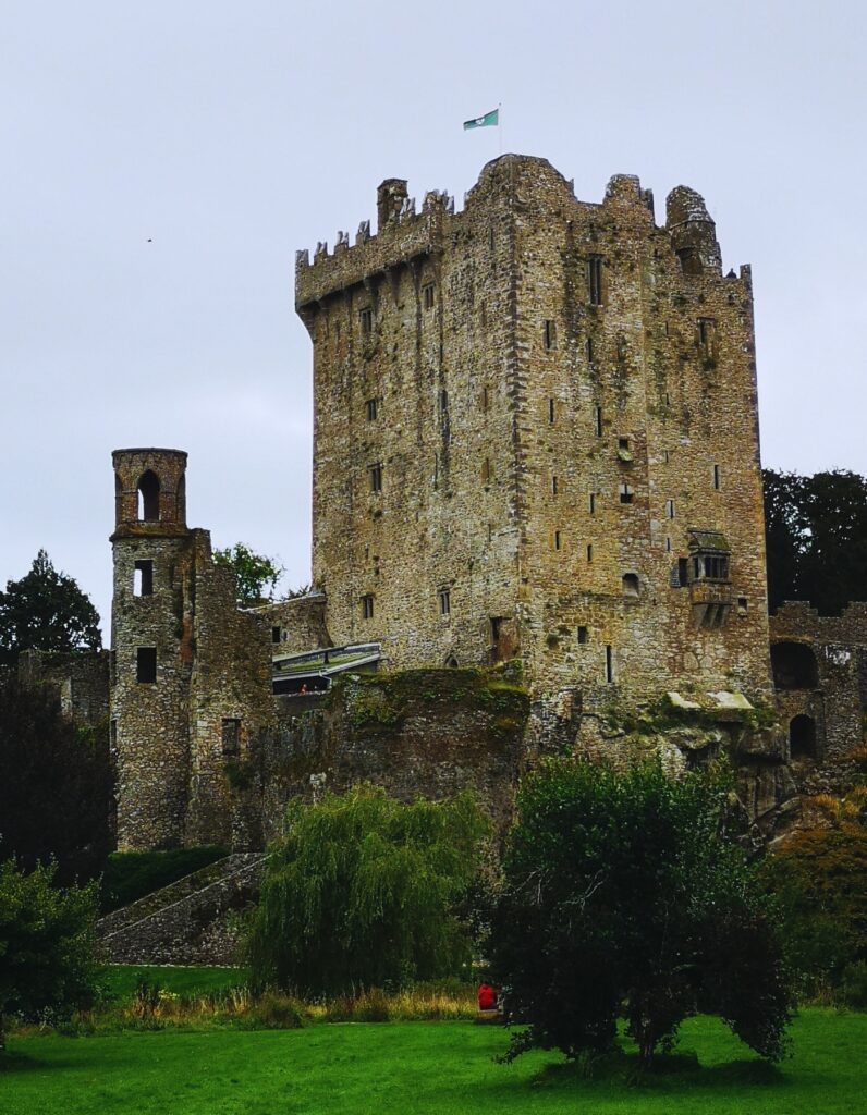 Blarney Castle in Ireland rises tall with its iconic medieval stone tower, surrounded by lush greenery and a cloudy gray sky. A small flag waves at the top, and visitors can be seen near the famous Blarney Stone platform.