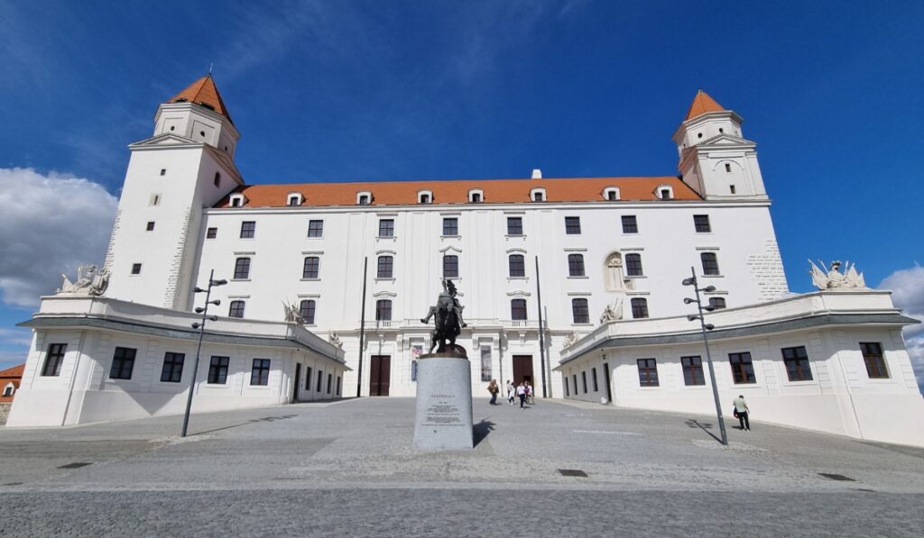 Front-facing shot of Bratislava Castle with its symmetrical white facade and four corner towers, a central statue of Svatopluk I on horseback in the courtyard, and a few visitors walking up the cobbled path.