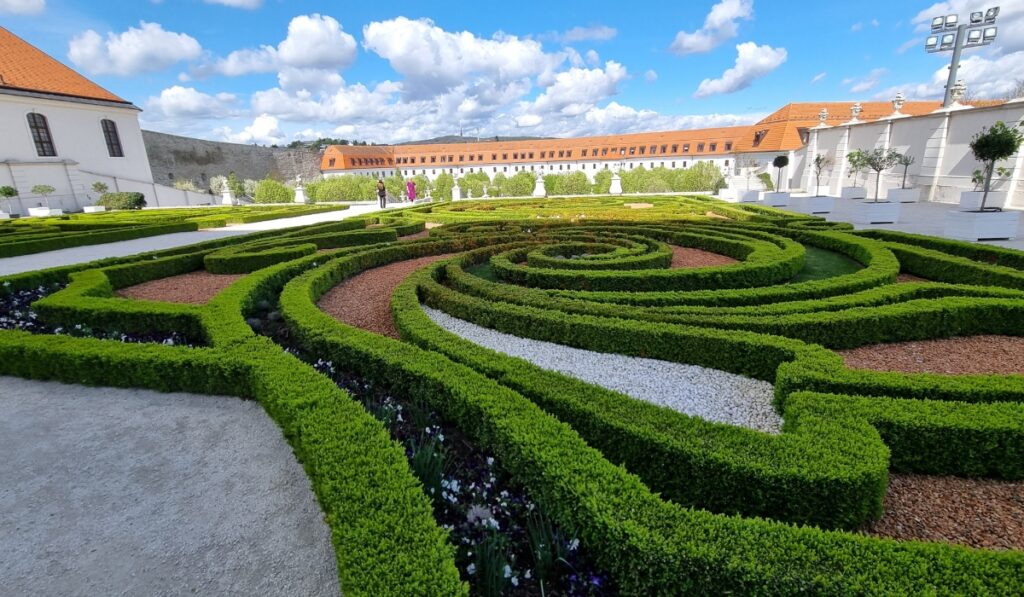 Intricately designed garden with spiral and geometric boxwood hedges bordered by gravel and mulch paths, set against the bright white walls and terracotta rooftops of Bratislava Castle grounds.