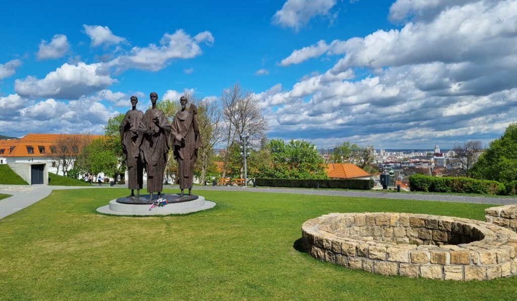 A bronze statue of three figures stands prominently in a manicured garden at Bratislava Castle, with a circular stone structure nearby and the city skyline in the background under a partly cloudy sky.