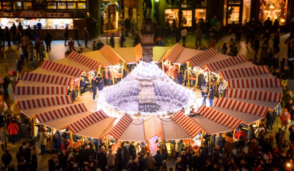 Overhead view of the Bratislava Christmas Market at night, with red-and-white striped stalls arranged in a circle around a fountain wrapped in sparkling fairy lights. The square is bustling with people enjoying food, drinks, and holiday cheer under the glow of warm lights.
