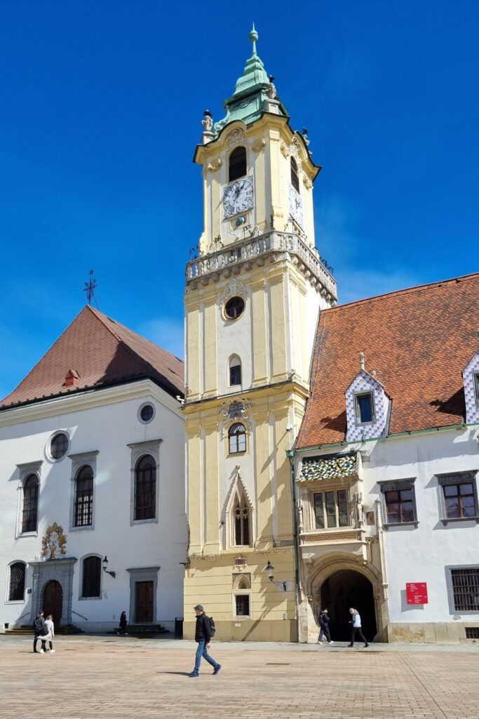 Old Town Hall in Bratislava, featuring a pastel yellow clock tower with ornate details and a green spire, surrounded by charming red-roofed buildings in the city’s main square.