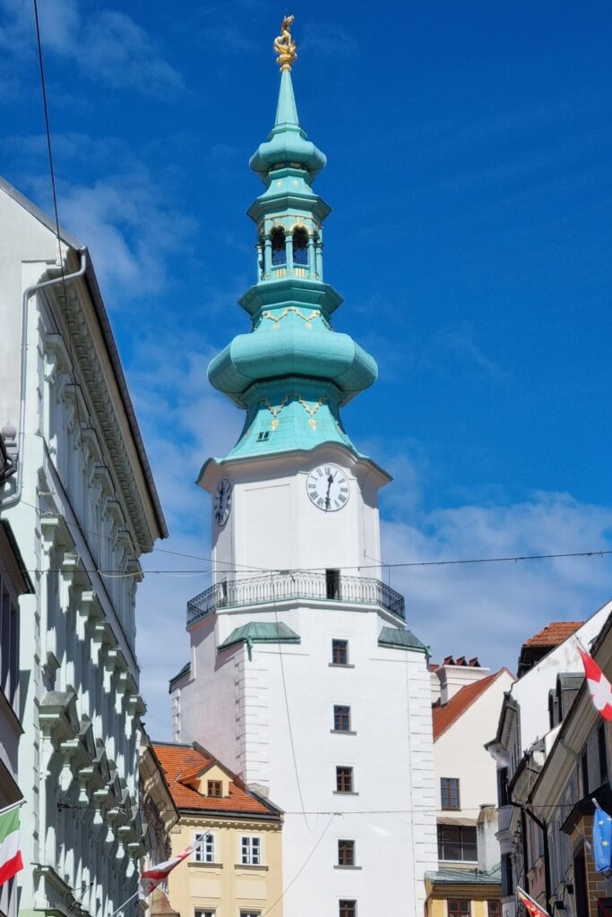 View of Michael’s Gate, a striking white medieval tower with a mint-green spire and golden ornament at the top, set against a bright blue sky and flanked by historic buildings.