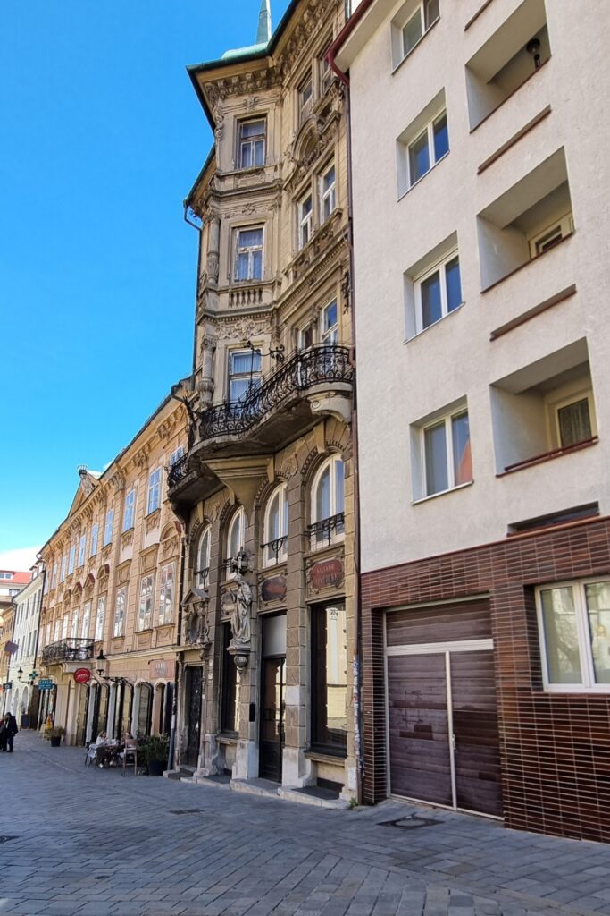 Historic buildings line a cobblestone street in Bratislava’s Old Town, with a baroque-style facade featuring statues and wrought-iron balconies alongside more modern architecture.