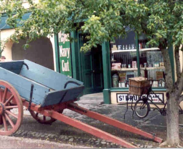 A vintage wooden cart with red wheels and a blue body rests beneath a leafy tree in front of a traditional shopfront at Bunratty Folk Park in Ireland. A classic bicycle with a woven basket leans nearby, and old-fashioned goods are visible through the shop window.