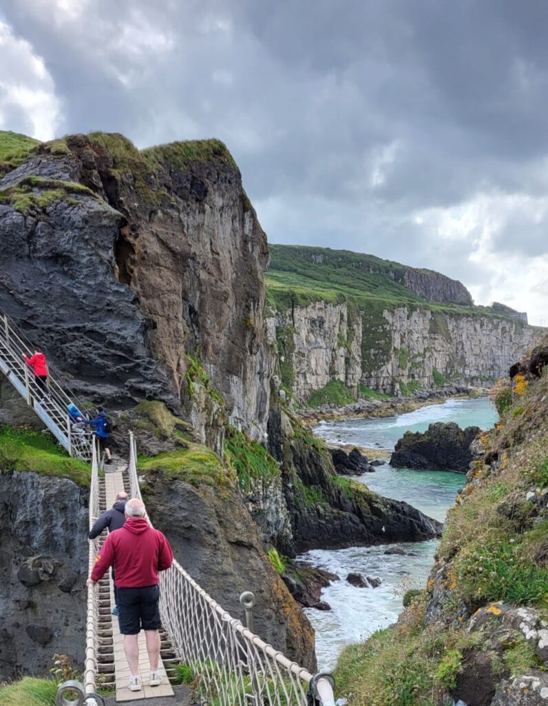 Tourists cross the Carrick-a-Rede Rope Bridge in Northern Ireland, suspended between steep rocky cliffs above turquoise coastal waters. Lush green grass covers the cliffs, while dramatic skies loom overhead, adding to the wild atmosphere of the scene.