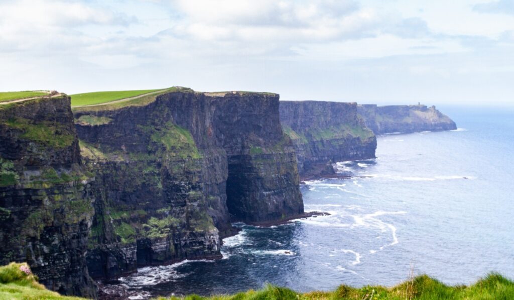 The dramatic Cliffs of Moher rise steeply from the Atlantic Ocean along Ireland’s west coast, with sheer rock faces covered in patches of green grass. Waves crash below while a distant tower sits atop the cliffs under a partly cloudy sky.