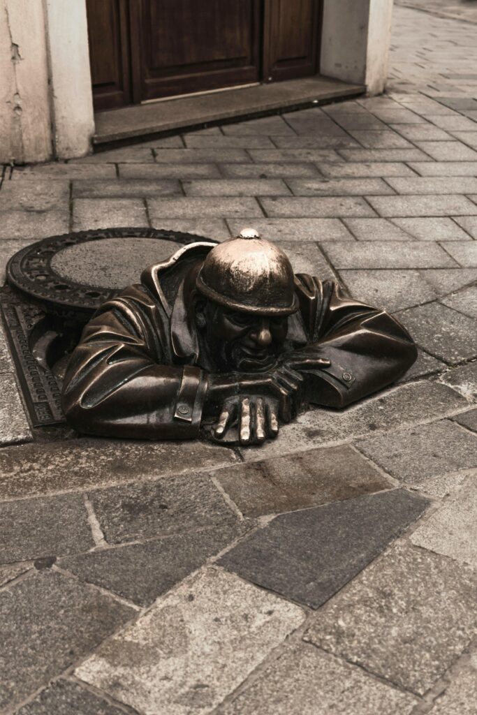 Bronze statue of a smiling man in a helmet emerging playfully from a manhole in a cobblestone street in Bratislava, known as "Čumil." His arms rest on the pavement as he peers out with a cheeky grin.