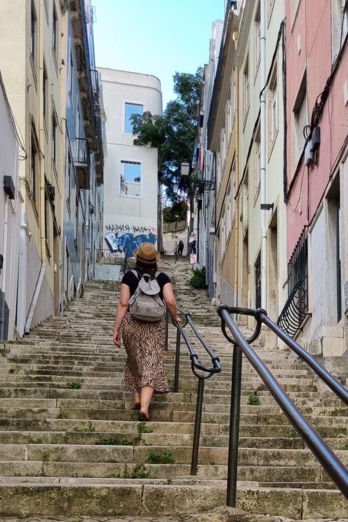 Me wearing a tan hat, black shirt, and zebra-print skirt walks up a long stone staircase between colorful, aged buildings. She carries a small light gray daypack and is surrounded by old railings and faint graffiti on the upper wall ahead, suggesting a European city, possibly Lisbon.