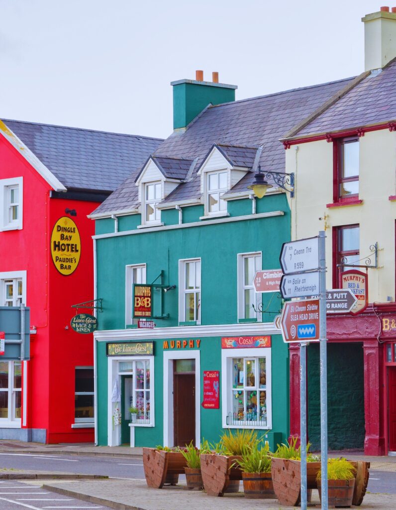 Colorful buildings in the town of Dingle, Ireland, including a bright red hotel, a green bed and breakfast, and a maroon pub. Shop signs and road signs point toward local attractions like Slea Head Drive, while flower planters line the sidewalk.