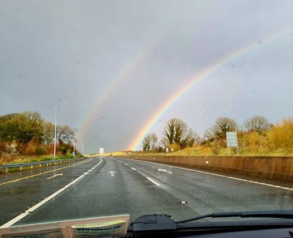 A double rainbow arches across a wet Irish highway, seen from inside a moving car. The sun breaks through stormy skies, lighting up the road and surrounding trees with a magical glow.