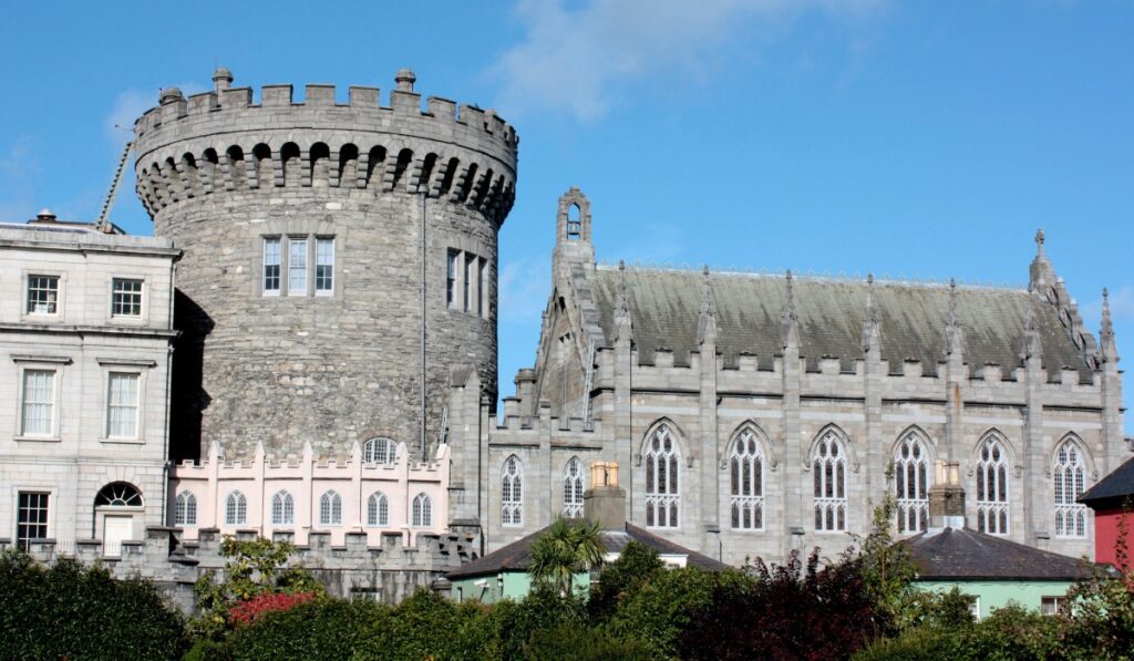 A view of Dublin Castle in Ireland, featuring its round medieval tower alongside a grand Gothic-style building with tall arched windows. The gray stone structures stand against a clear blue sky, surrounded by colorful buildings and greenery.