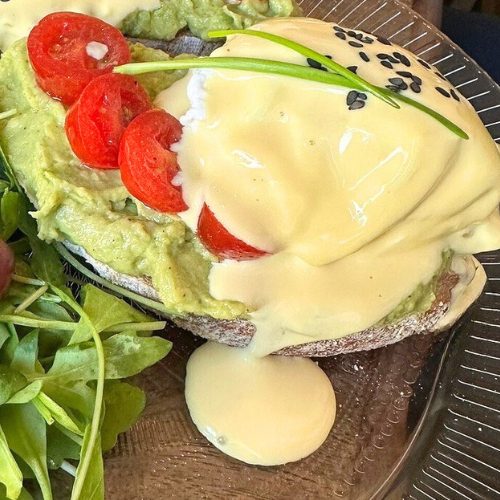 Sourdough toast topped with creamy avocado spread, halved cherry tomatoes, and a perfectly poached egg smothered in rich hollandaise sauce, garnished with black sesame seeds and a sprig of chive. A side of fresh arugula adds a crisp, peppery touch to the dish, served on a glass plate.
