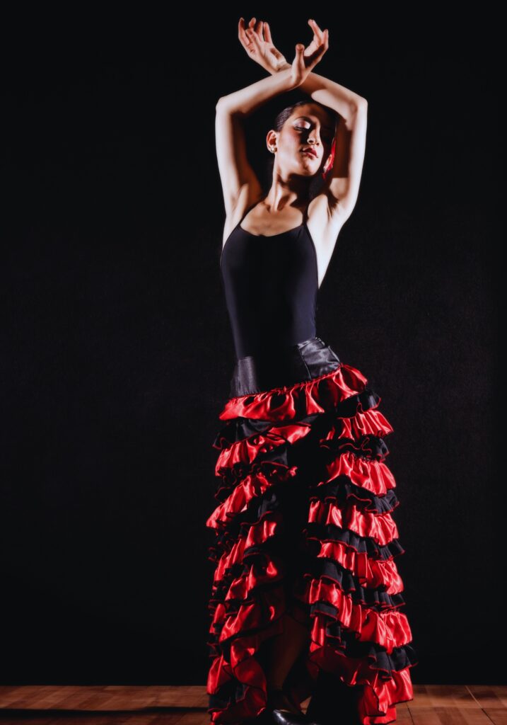 A flamenco dancer strikes a graceful pose against a dark background, wearing a black top and a ruffled red-and-black skirt. Her arms are elegantly raised above her head, capturing the passion and poise of traditional Spanish dance.