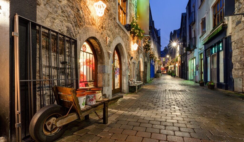A cozy cobblestone street in Galway City, Ireland, lit by warm string lights and shop windows at dusk. Rustic stone buildings with arched doorways are decorated with festive garlands, and a wooden cart with painted flower pots rests by a storefront.