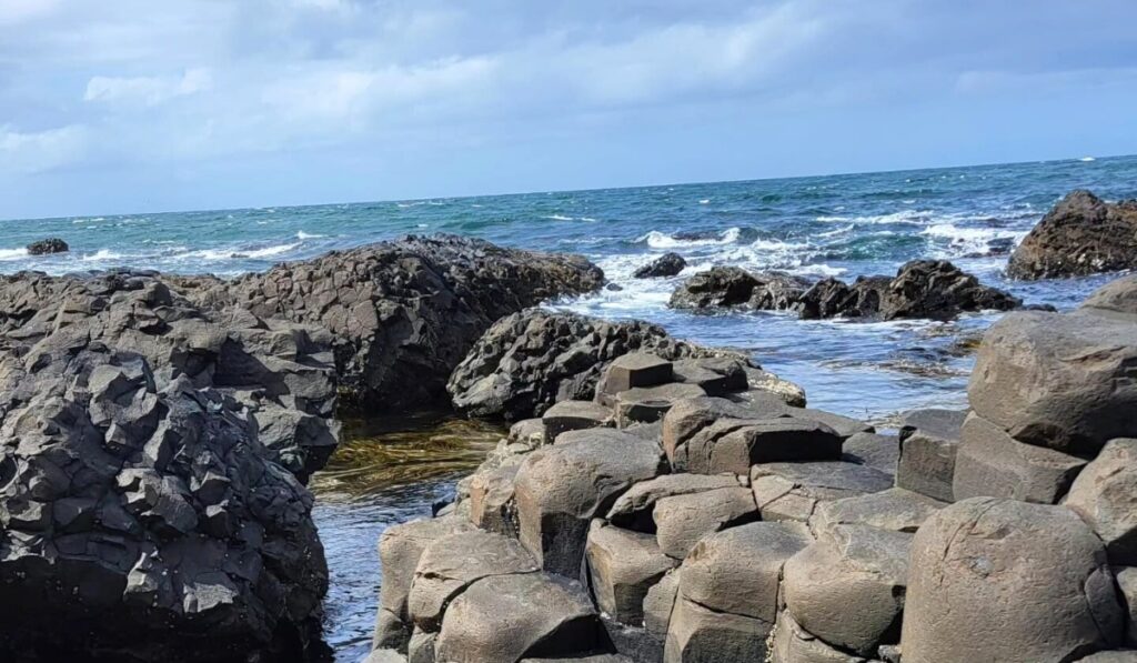 The Giant’s Causeway in Northern Ireland features interlocking basalt columns along a rugged coastline, with ocean waves crashing against the dark volcanic rock formations. The scene is bright and breezy under a partly cloudy sky.