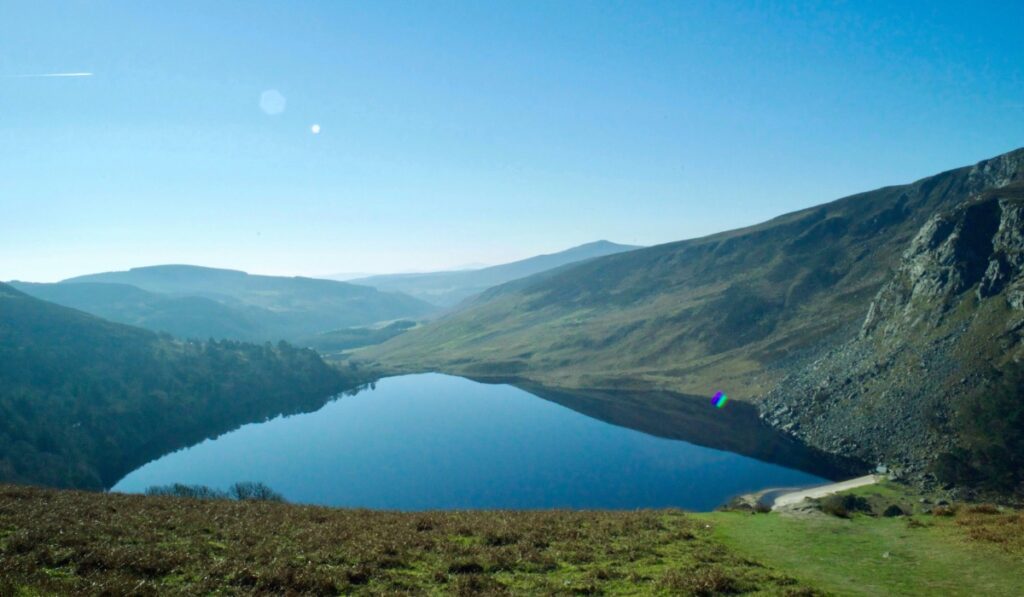 A serene view of Glendalough in Ireland, showcasing a perfectly still lake reflecting the surrounding green hills and rugged cliffs under a clear blue sky. The landscape stretches into distant misty mountains, giving a peaceful, untouched vibe.