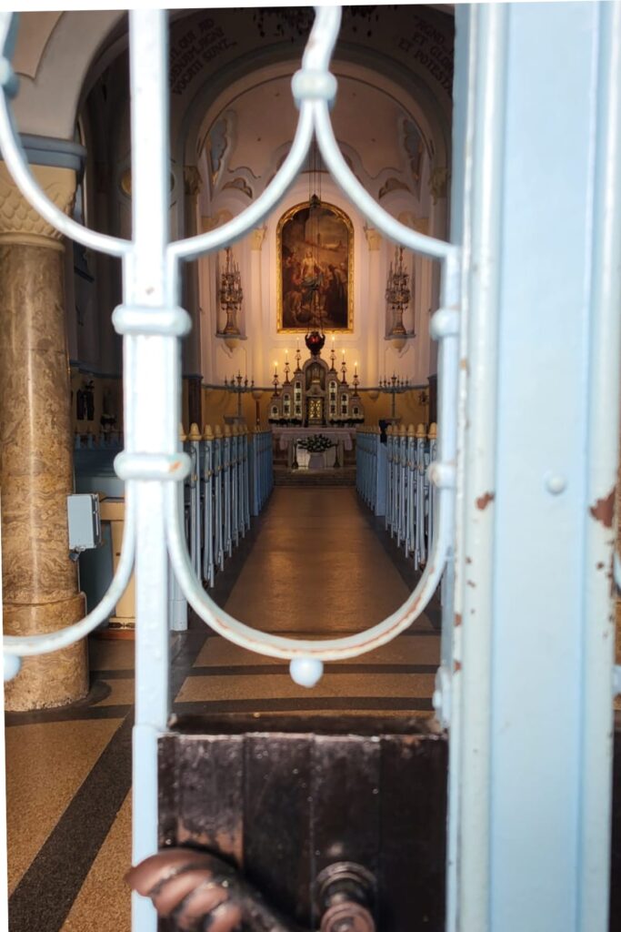 Looking through ornate iron bars into the interior of the Blue Church in Bratislava, revealing a softly lit altar, light blue pews, and religious artwork under an arched ceiling.